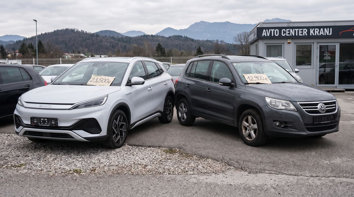 Dva rabljena avtomobila na dvorišču prodajalca — kitajski EV in starejši evropski SUV s cenikoma na vetrobranskem steklu